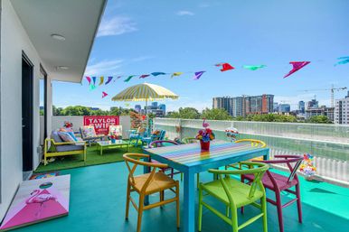Vibrant rooftop patio with a rainbow-striped dining table and mismatched colorful chairs, yellow-striped umbrella, string of multicolored pennant flags, lounge seating on artificial turf, flamingo cornhole board, potted flowers and an urban skyline under a bright blue sky.