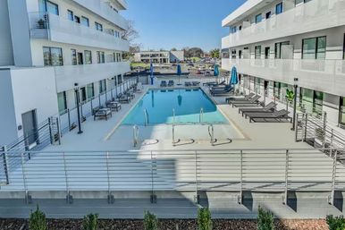 Sunlit modern white apartment courtyard with a central rectangular outdoor pool, sleek lounge chairs and blue umbrellas on a raised deck, balconies overlooking the pool and an urban parking lot beyond.