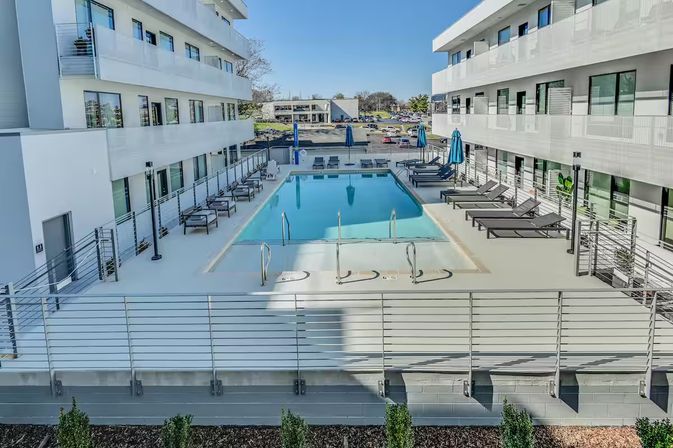 Sunlit modern white apartment courtyard with a central rectangular outdoor pool, sleek lounge chairs and blue umbrellas on a raised deck, balconies overlooking the pool and an urban parking lot beyond.