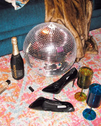 Glam home party still life on a colorful rug: mirrored disco ball, black patent stiletto heels, bottle of sparkling wine, faux-fur throw and tinted goblets.