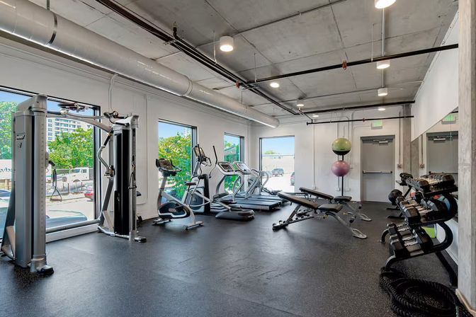 Sunlit modern fitness center in an urban building with large windows, treadmills and ellipticals, a cable machine, adjustable bench, free weights and stability balls on a black rubber floor.