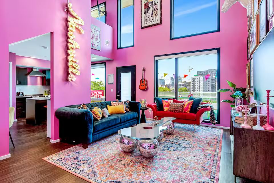 Vibrant pink modern loft living room with high ceilings and large windows overlooking a city skyline, navy velvet sofa and red loveseat with colorful pillows, chrome orb coffee table on a patterned rug, wall-mounted guitar and open kitchen visible.