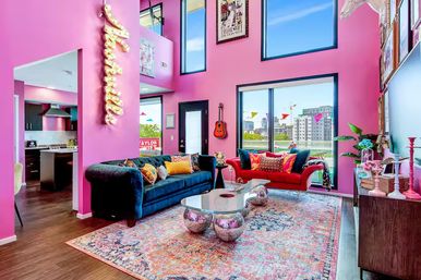 Vibrant pink modern loft living room with high ceilings and large windows overlooking a city skyline, navy velvet sofa and red loveseat with colorful pillows, chrome orb coffee table on a patterned rug, wall-mounted guitar and open kitchen visible.