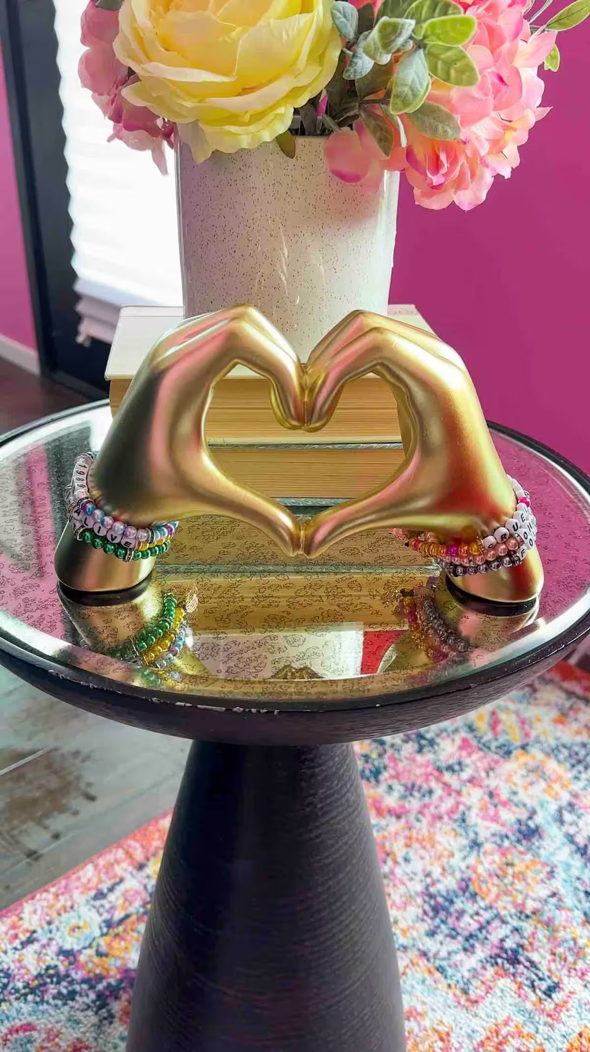 Gold hand sculpture forming a heart on a round glass side table with colorful beaded bracelets, white speckled vase of pink and yellow flowers behind, bright magenta wall and multicolored rug in the background.