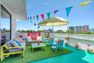 Festive colorful urban rooftop patio with striped yellow umbrella, turquoise chairs, bunting flags, artificial grass and downtown skyline view.