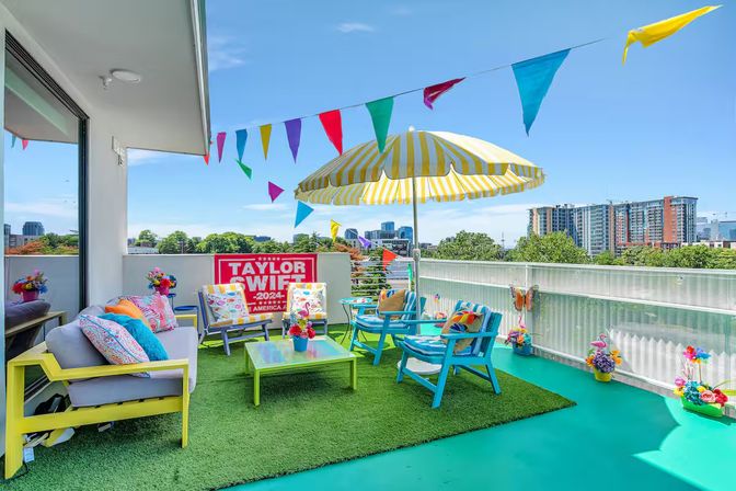 Festive colorful urban rooftop patio with striped yellow umbrella, turquoise chairs, bunting flags, artificial grass and downtown skyline view.