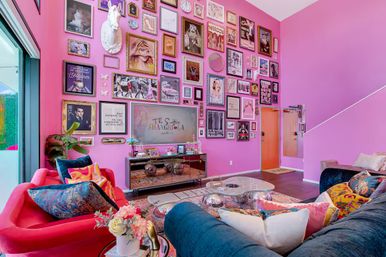 Bright pink living room with a towering gallery wall of framed art and a white unicorn bust, colorful velvet sofas with patterned pillows, mirrored console and glass coffee table in a playful, eclectic home interior.