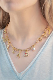 Close-up of gold paperclip chain charm necklace on a person's neck, playful charms including key, bee, pearl drop, crescent moon, wishbone and cherries, paired with a light blue V-neck tee