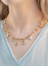 Close-up of gold paperclip chain charm necklace on a person's neck, playful charms including key, bee, pearl drop, crescent moon, wishbone and cherries, paired with a light blue V-neck tee