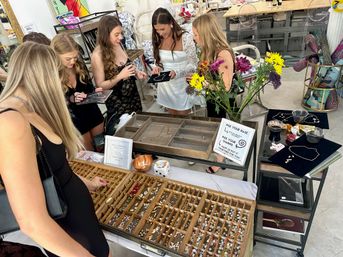 Women in party dresses choosing charms and necklaces at a bright boutique DIY jewelry bar, wooden charm tray, velvet necklace displays and a vase of fresh flowers on the table.