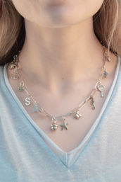 Close-up of a silver paperclip-chain charm necklace on a light blue V-neck top, featuring charms including a starfish, seahorse, acorn, sun, anchor, bee, wine glass and letter S.