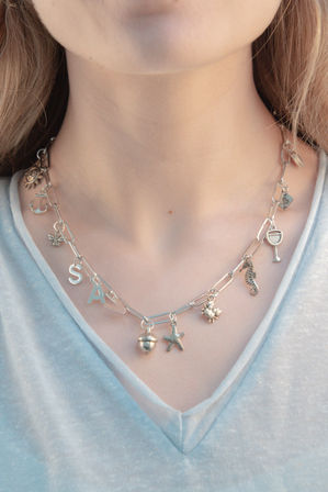 Close-up of a silver paperclip-chain charm necklace on a light blue V-neck top, featuring charms including a starfish, seahorse, acorn, sun, anchor, bee, wine glass and letter S.