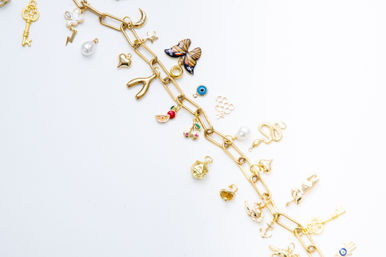 Flat-lay gold charm bracelet on a white background, diagonal chain studded with playful charms: enamel butterfly, wishbone, snake, vintage key, pearl drop, evil eye, cherry and heart accents.