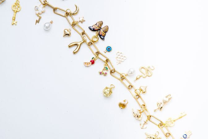 Flat-lay gold charm bracelet on a white background, diagonal chain studded with playful charms: enamel butterfly, wishbone, snake, vintage key, pearl drop, evil eye, cherry and heart accents.