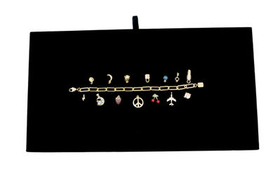 Gold charm bracelet with assorted miniature charms (peace sign, airplane, cherries, moon, lock and more) neatly displayed on a black velvet jewelry board — chic jewelry flat lay