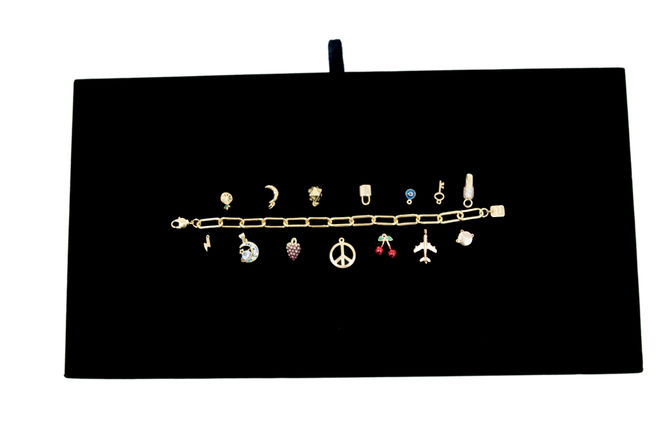Gold charm bracelet with assorted miniature charms (peace sign, airplane, cherries, moon, lock and more) neatly displayed on a black velvet jewelry board — chic jewelry flat lay