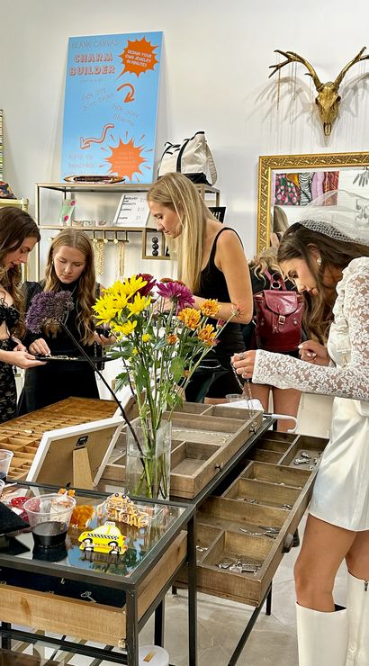 Four women browsing necklaces at a bright jewelry boutique table with wooden display trays, a vase of sunflowers, a “Charm Builder” sign, and one woman in a white lace bridal outfit.
