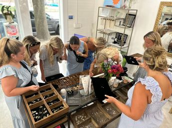 Group of women shopping and examining rings, earrings and necklaces arranged in wooden trays on a bright boutique counter with a small flower bouquet.