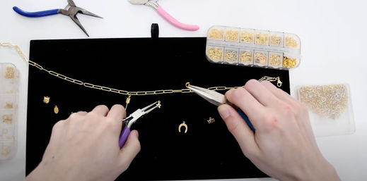 Hands using pliers to attach gold charms to a paper‑clip chain necklace on a black work mat, surrounded by containers of gold jump rings and jewelry tools — DIY jewelry making scene.