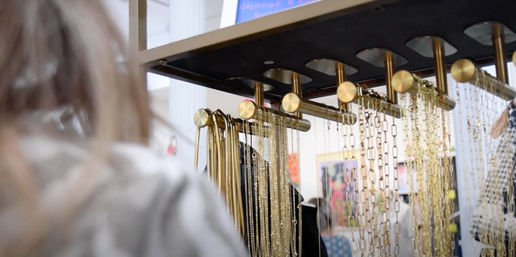 Hanging gold necklaces and chains on brass display rods in an indoor boutique jewelry display, with a blurred shopper in the foreground and colorful artwork in the background.