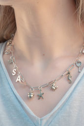 Close-up of a neck wearing a silver charm chain with initial letter charms and seaside-themed pendants (starfish, seahorse, crab, acorn and cocktail glass) over a pale blue V-neck top.