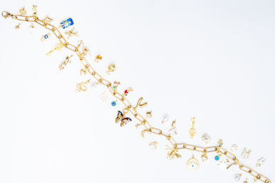Gold charm bracelet flat-lay on white background, playful fashion jewelry with assorted enamel, pearl, and gemstone charms — butterfly, evil eye, keys, moon and star.