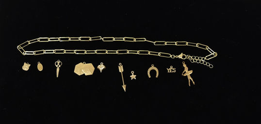 Gold chain charm necklace flat-lay on black background featuring playful gold charms: cat face, oval tag, scissors, geometric tags, heart, arrow, star, horseshoe, "YES" word and ballerina pendant