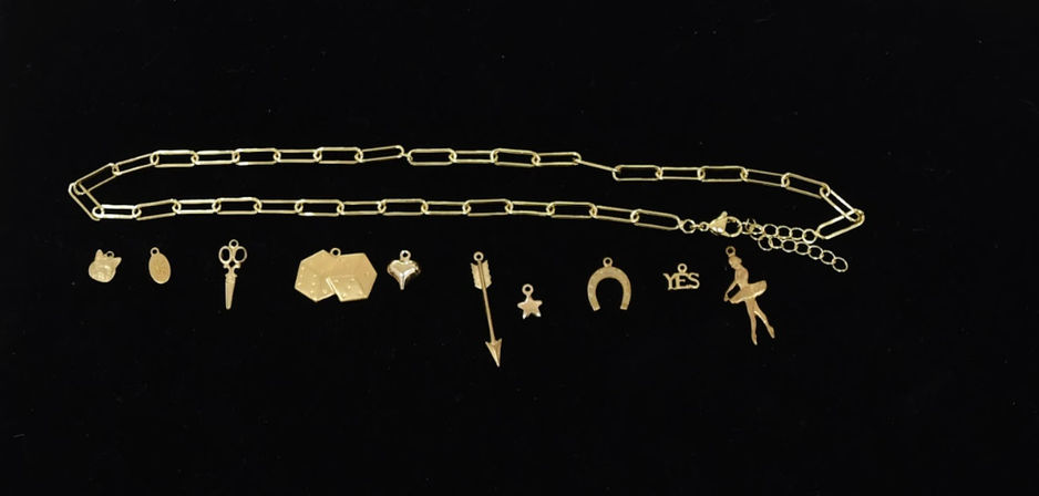 Gold chain charm necklace flat-lay on black background featuring playful gold charms: cat face, oval tag, scissors, geometric tags, heart, arrow, star, horseshoe, "YES" word and ballerina pendant