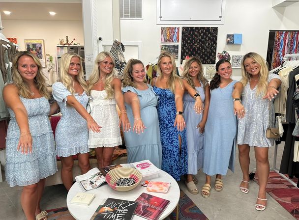 Eight smiling women in light blue and white summer dresses lined up inside a clothing boutique, extending their hands to show rings; clothing racks, colorful textiles and a round display table with magazines in the foreground.