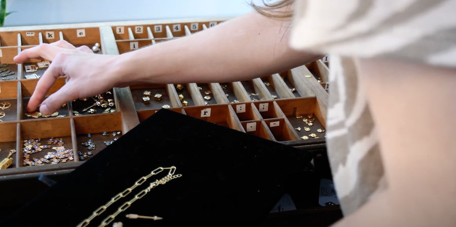 Hand selecting gold jewelry findings from a wooden divided tray beside a black pad with a gold chain in an artisan jewelry workshop
