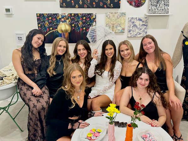 Eight women in stylish black and white outfits pose inside a cozy art-filled boutique; the woman in a white lace dress holds a lace parasol while friends smile around a table with flowers and treats.
