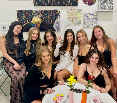 Eight women in stylish black and white outfits pose inside a cozy art-filled boutique; the woman in a white lace dress holds a lace parasol while friends smile around a table with flowers and treats.