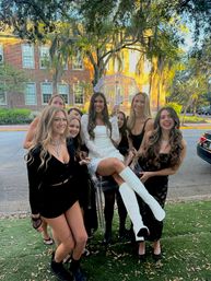 Smiling group of young women posing outdoors; one in a white mini dress and knee-high white boots sits on a clear chair holding a lace parasol, historic brick building and oak trees draped with Spanish moss in warm evening light.
