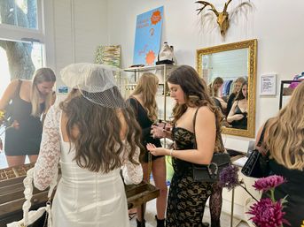 Bridal party shopping in a trendy fashion boutique — bride-to-be in white satin dress with birdcage veil examines jewelry with friends near a gold-framed mirror and decorative wall antlers.