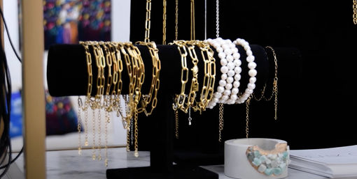 Black velvet T-bar jewelry display with gleaming gold chain bracelets and stacked white pearl bracelets, beside a concrete dish of aqua gemstone beads on a marble countertop in a boutique-style setting.