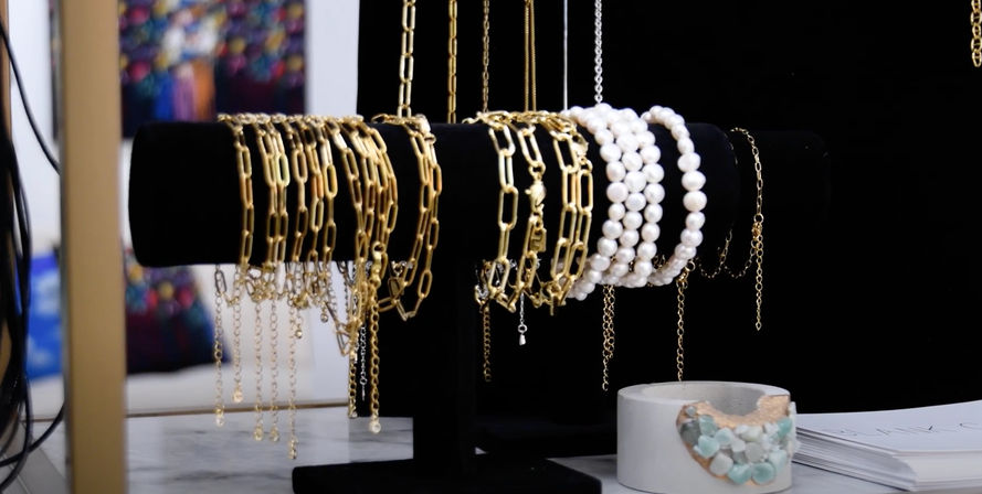 Black velvet T-bar jewelry display with gleaming gold chain bracelets and stacked white pearl bracelets, beside a concrete dish of aqua gemstone beads on a marble countertop in a boutique-style setting.