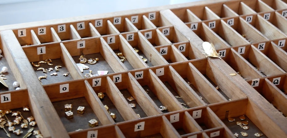 Mini treasure trove: wooden divided tray with numbered and lettered slots filled with tiny gold-tone charms and jewelry findings on a craft workshop bench