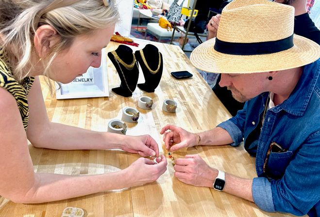 Two shoppers at an indoor craft fair lean over a wooden table inspecting gold-tone necklaces and bracelets on black busts and small display bowls; one wears a straw hat and denim shirt.