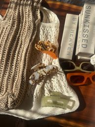 Cozy sunlit flatlay on a wooden table with beige and cream knitwear, neutral-toned boxes, tortoiseshell and amber hair claw clips, a translucent pale‑green clip, and olive and orange rectangular sunglasses.