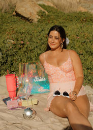 Smiling bride-to-be relaxing on a sunny beach blanket by coastal dunes, iridescent "Bride" tote, pink floral top, white sunglasses and picnic items nearby