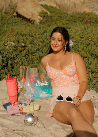 Smiling woman relaxing on a beach blanket in golden-hour light, wearing a pink floral crop top and skirt and holding white sunglasses; iridescent tote labeled "Bride", pink tumbler, disco-ball cup and small beauty items arranged beside her amid coastal greenery.