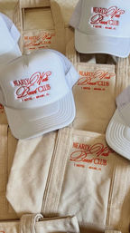 Stack of white trucker hats and beige canvas tote bags embroidered with a red beach-club logo and small Miami, FL hotel text — casual coastal merchandise.