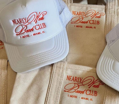Stack of white trucker hats and beige canvas tote bags embroidered with a red beach-club logo and small Miami, FL hotel text — casual coastal merchandise.