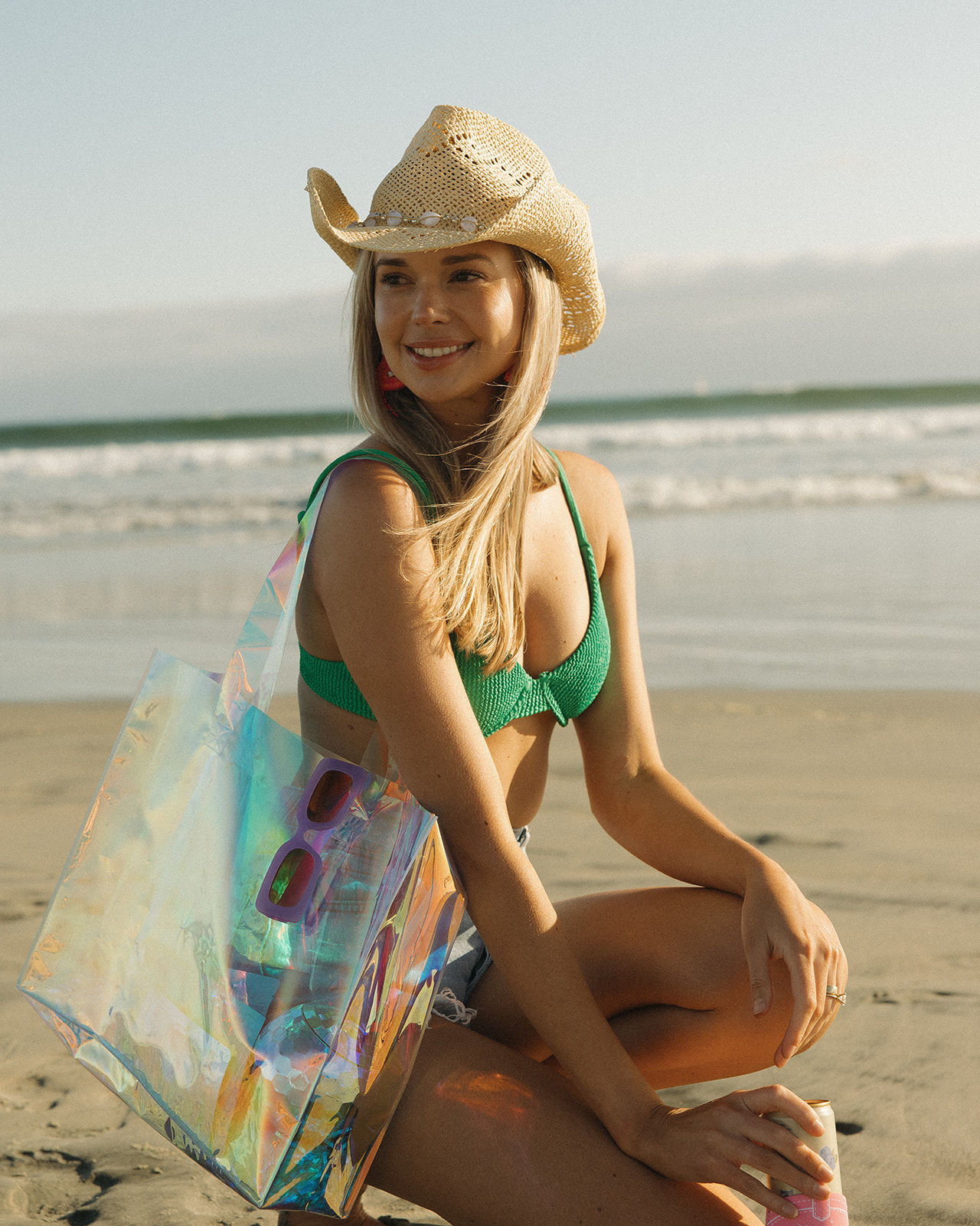 Smiling woman in a straw cowboy hat and green bikini on a sunny sandy beach with ocean waves, carrying a holographic tote and holding a canned drink.