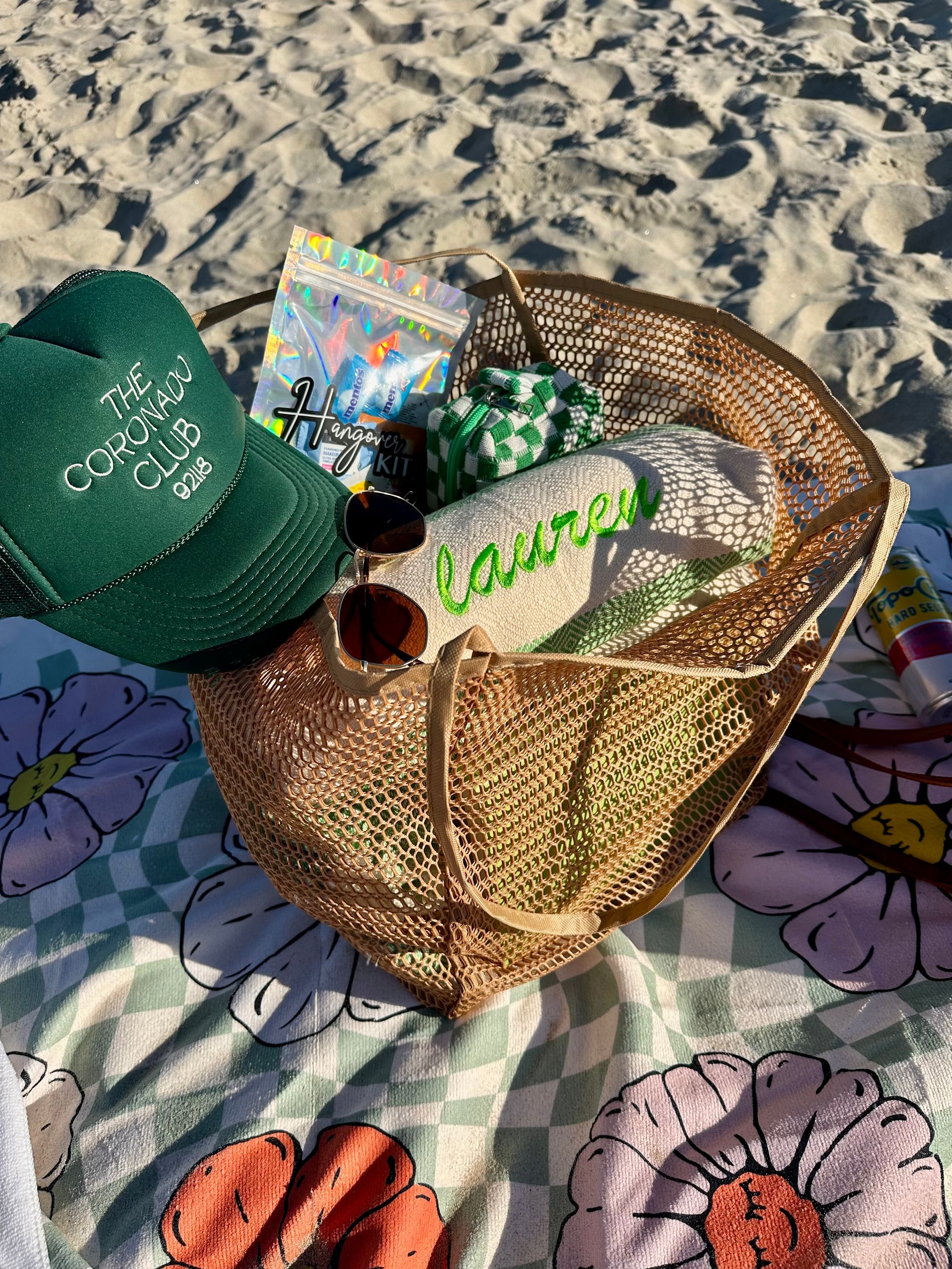 Mesh beach tote on a sunny sandy beach filled with beach essentials — sunglasses, holographic pouch, green checkered pouch, personalized embroidered towel and a green trucker hat resting on a floral blanket.