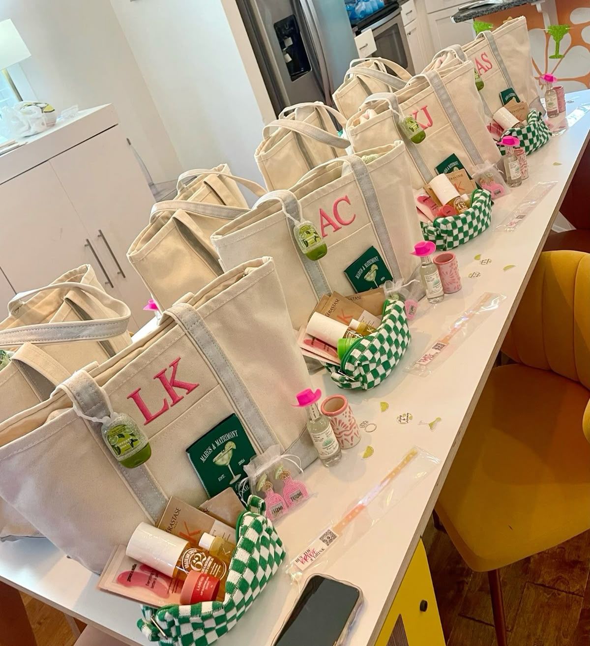 Personalized canvas tote gift bags with monogram initials, mini skincare items and green-check pouches neatly lined up on a bright kitchen island as party favors