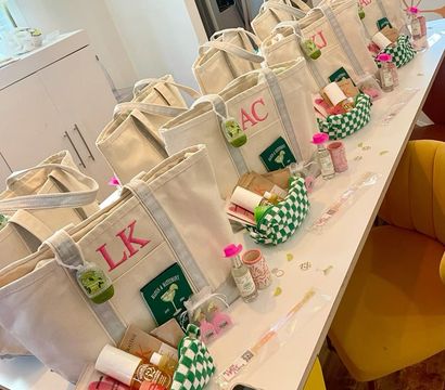 Personalized canvas tote gift bags with monogram initials, mini skincare items and green-check pouches neatly lined up on a bright kitchen island as party favors