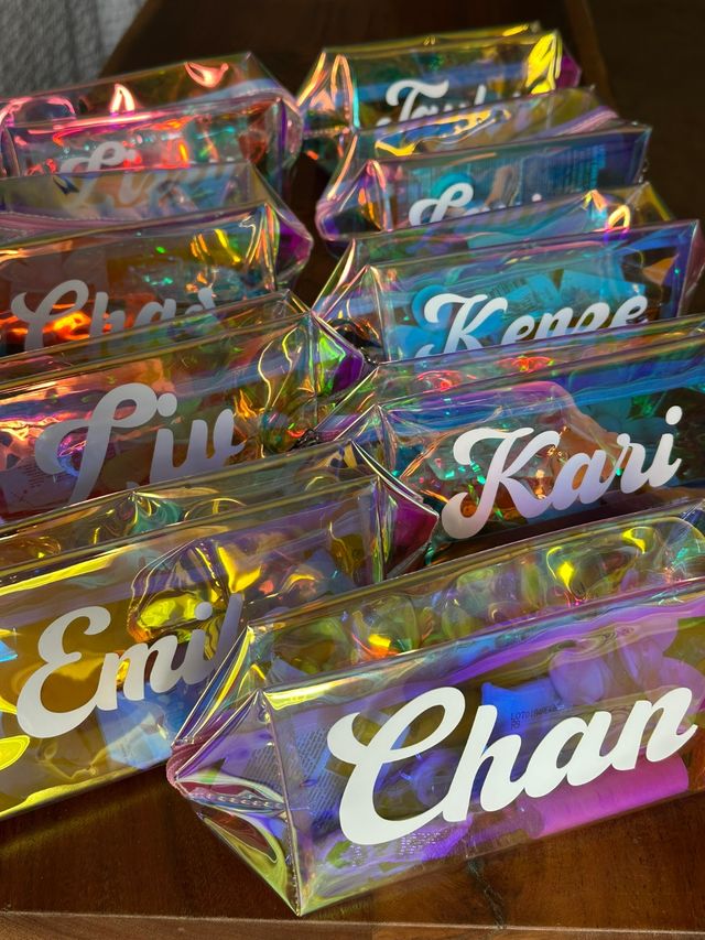 Iridescent holographic personalized pouches with white cursive name decals arranged on a wooden table, colorful rainbow reflections — custom pencil or makeup bags ideal for party favors