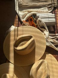 Sunlit flat lay of three wide-brim straw hats on a wooden table with cream knit scarves, two retro sunglasses, a tortoiseshell hair clip and stacked sunscreen boxes — summer beach accessories.