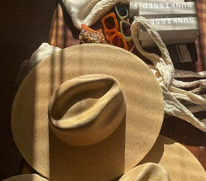 Sunlit flat lay of three wide-brim straw hats on a wooden table with cream knit scarves, two retro sunglasses, a tortoiseshell hair clip and stacked sunscreen boxes — summer beach accessories.